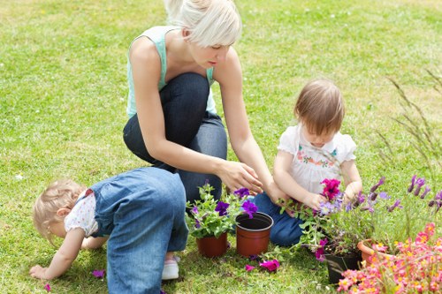 In a lush, well-maintained garden, an adult woman with short blonde hair, wearing a light blue tank top and dark jeans, crouches on a vibrant green lawn engaging in gardening activities. She is seen tending to flowering plants in small pots, including bright pink petunias and purple lavender, set among a variety of other colorful blooms. Two young children, a girl with blonde hair in a white floral dress and a boy in a blue outfit, are also involved; one child is bent over, closely examining or picking flowers, while the other sits nearby, holding a small plant. The garden features a neatly trimmed hedge line in the background, creating a natural boundary, with a mix of flower beds and open grassy spaces. The scene appears to be in bright, natural daylight, possibly during a warm weather day, reflecting a peaceful outdoor environment suitable for family gardening activities. This image aligns with services provided by Gardening Services Edmonton, focusing on garden planting, maintenance, and outdoor care in the local Edmonton area, with a backdrop of fresh, natural textures and vibrant plant colours, highlighting the importance of outdoor landscaping and gardening expertise.