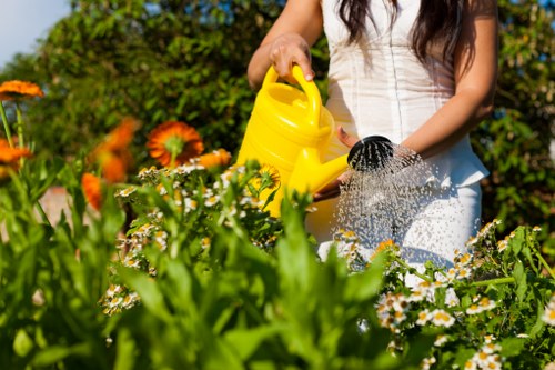 A garden scene featuring a well-maintained, lush green lawn area bordered by a variety of flowering plants, including orange and white blooms, with a background of dense foliage and hedges. In the foreground, a person wearing a white top is watering the garden using a yellow watering can, with water pouring onto the plants. The garden surface includes a paved pathway and areas of soil, indicating attentive landscaping and outdoor maintenance. The scene is illuminated by natural daylight, suggesting pleasant weather, and reflects a professionally cared-for outdoor space typical of residential gardens in Edmonton, aligning with gardening services provided by Gardening Services Edmonton.