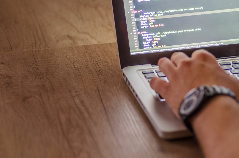 A person's hands working on a laptop with a dark screen displaying lines of colorful code in a text editor. The laptop is placed on a reflective black surface, and the background is blurred but sugges
